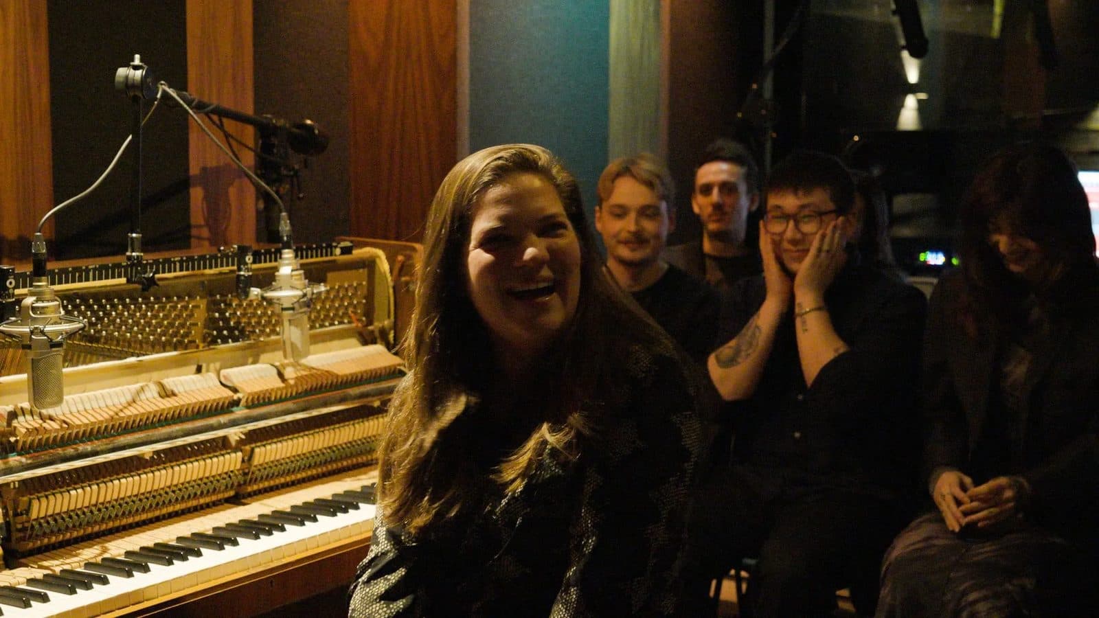 Host smiling beside the studio piano with the audience close behind