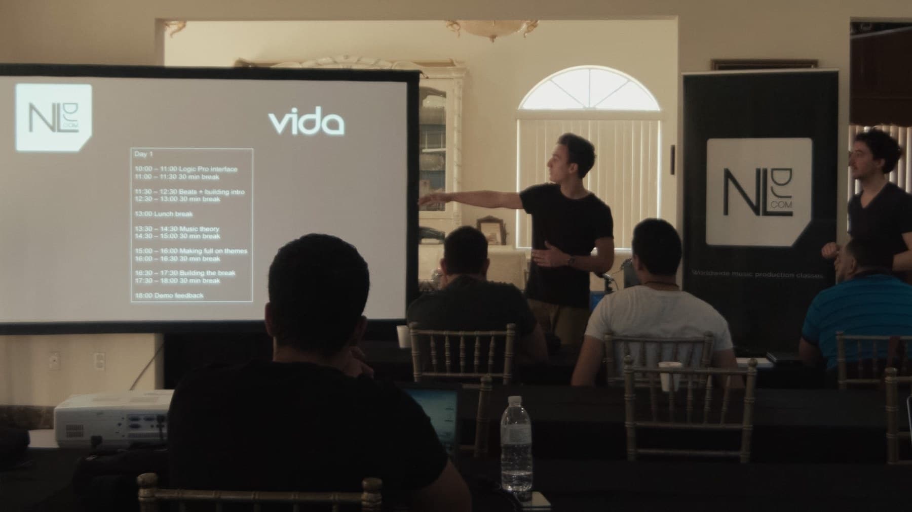 Ivo Adelaar teaching a production masterclass in Miami, pointing at a projected day schedule under the Vida and NLDJ logos, students seated at laptops