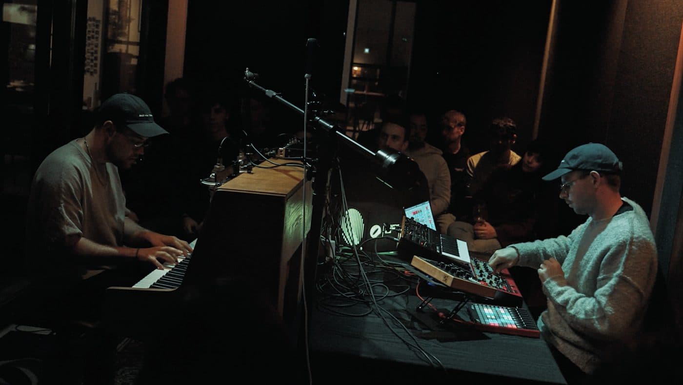 Two artists playing an opened upright piano and a synth rig side by side during a Disruptive Records session, the audience in the dark